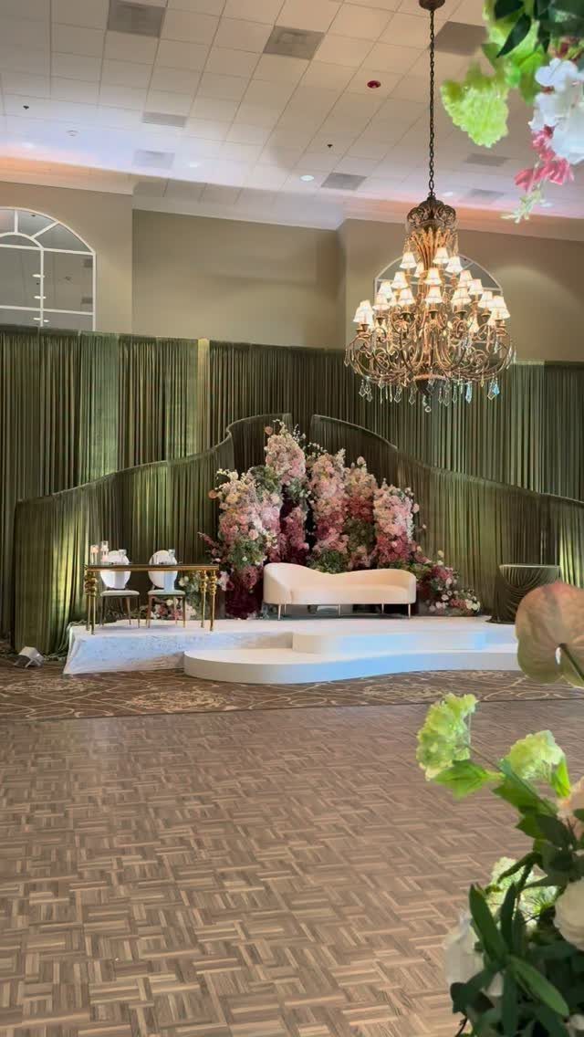 Wedding stage with pink and purple flowers against olive green curtains and crystal chandelier.