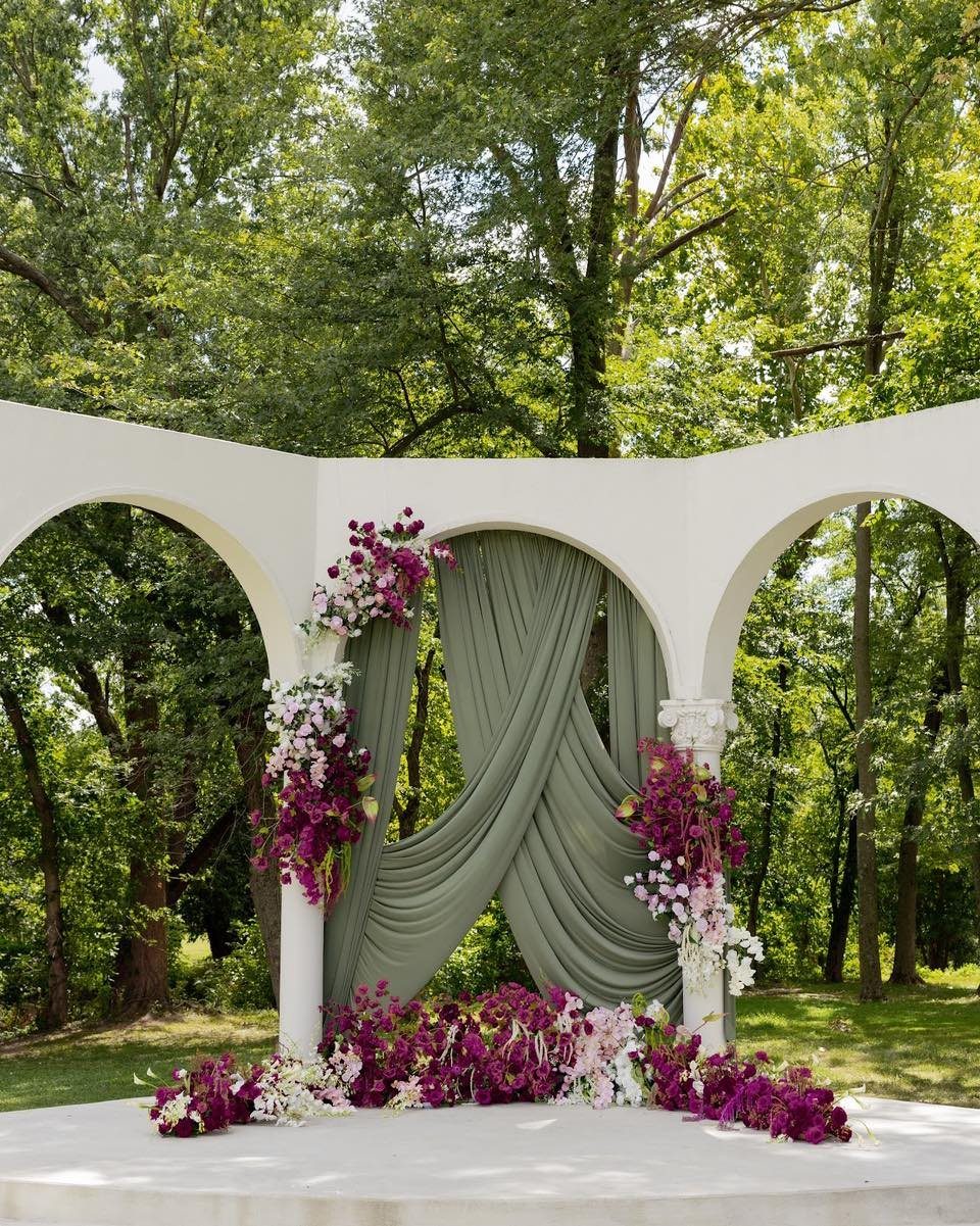 White arched gazebo with sage green drapery and purple and white floral arrangements.