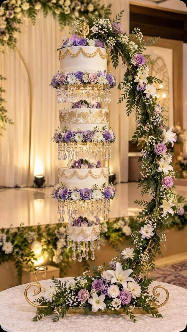 Multi-tiered white wedding cake with purple roses and gold trim under a floral arch.
