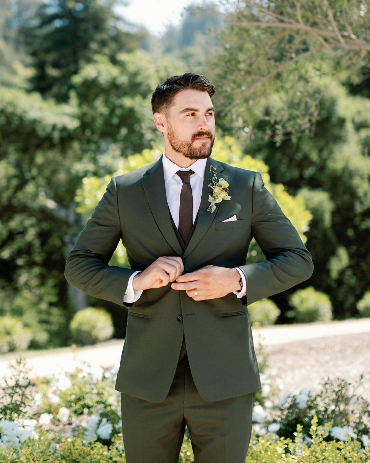 Man in a dark green wedding suit standing outdoors against a lush green background.