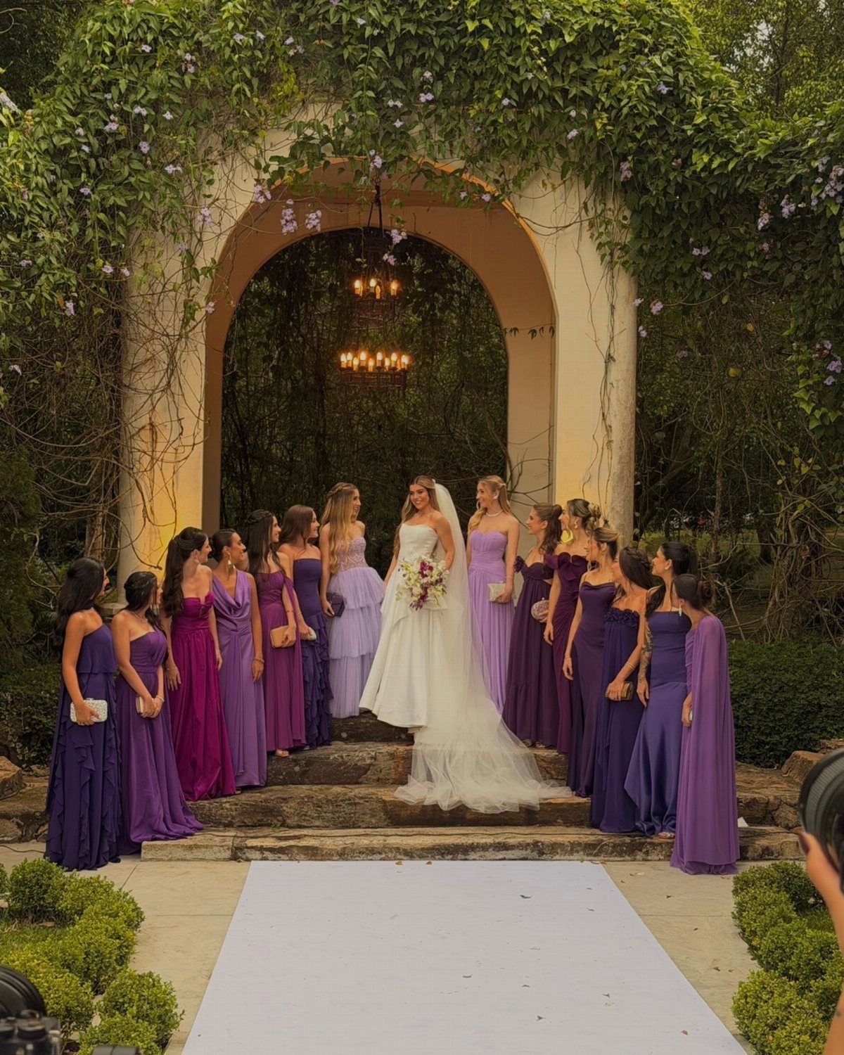 Bride in white gown surrounded by bridesmaids wearing various shades of purple dresses.