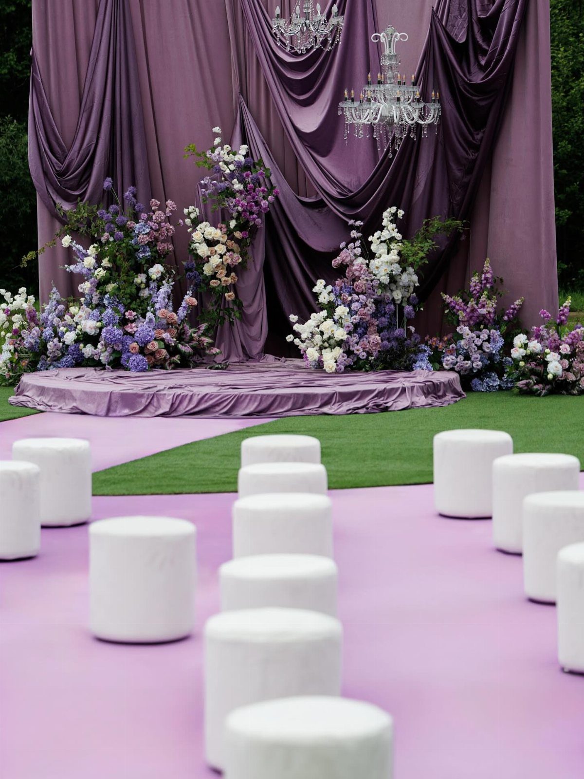 Purple draped backdrop with floral arrangements and white stools for a wedding ceremony.