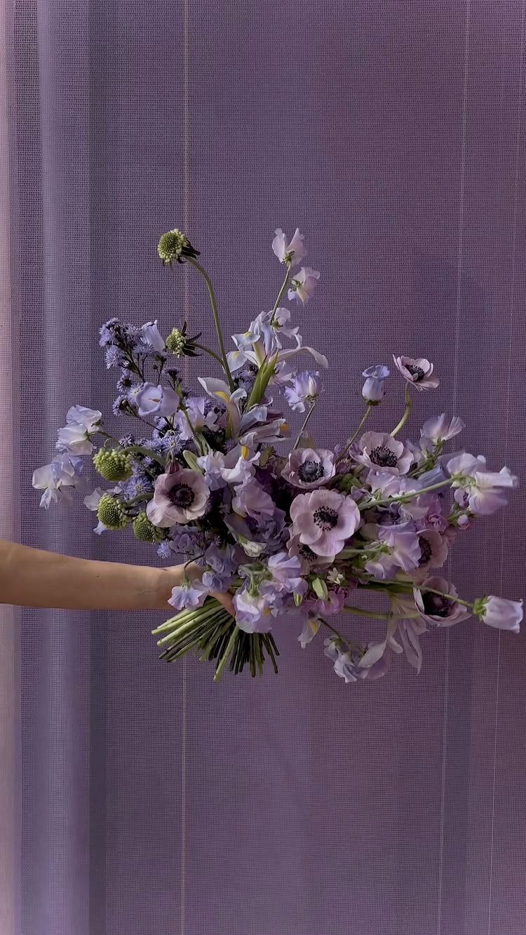 Purple and lavender flower bouquet with green stems held against purple fabric.