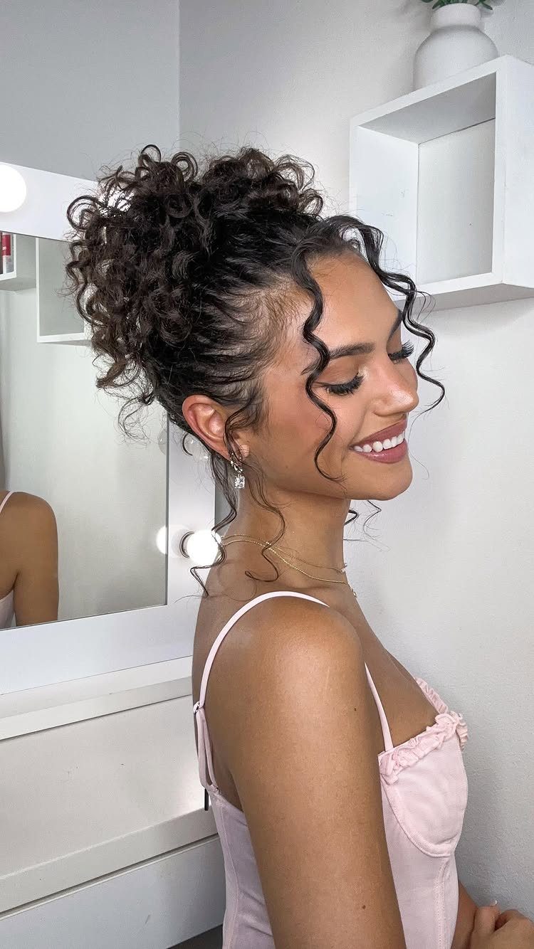18 Hoco Updo Hairstyles From Braids to Sleek Buns 5 A smiling young woman with dark curly hair styled in a voluminous updo with face-framing curls.