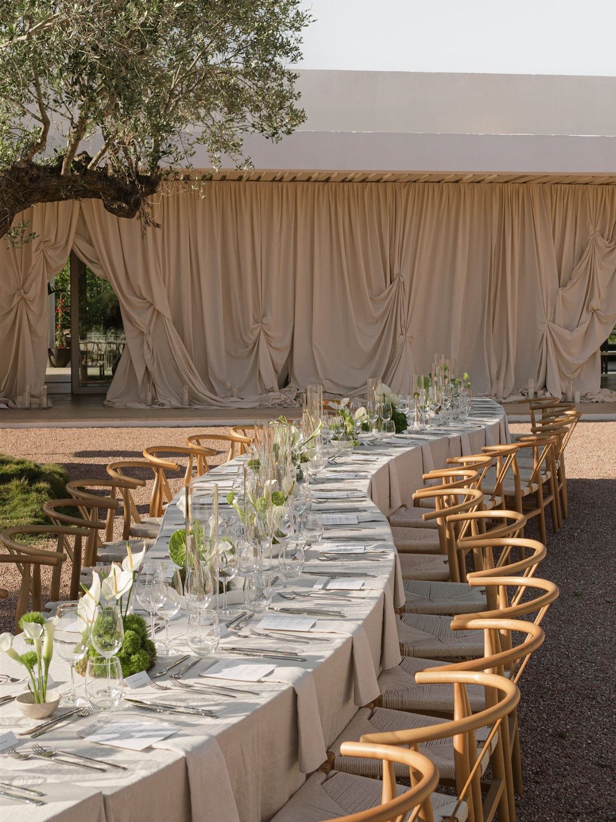 Curved wedding table with ivory linen tablecloth, glassware, and wooden chairs outdoors.