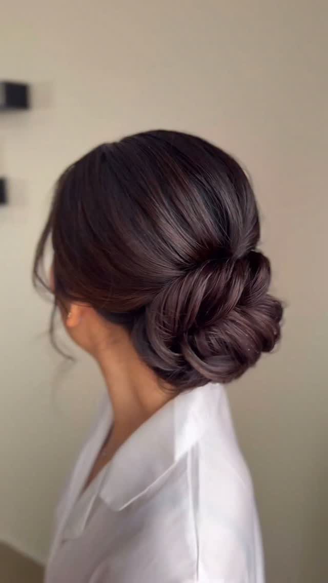18 Low Messy Bun for Wedding Ideas with Braids and Florals 4 Woman with dark brown hair styled in a polished, voluminous low bridal bun.