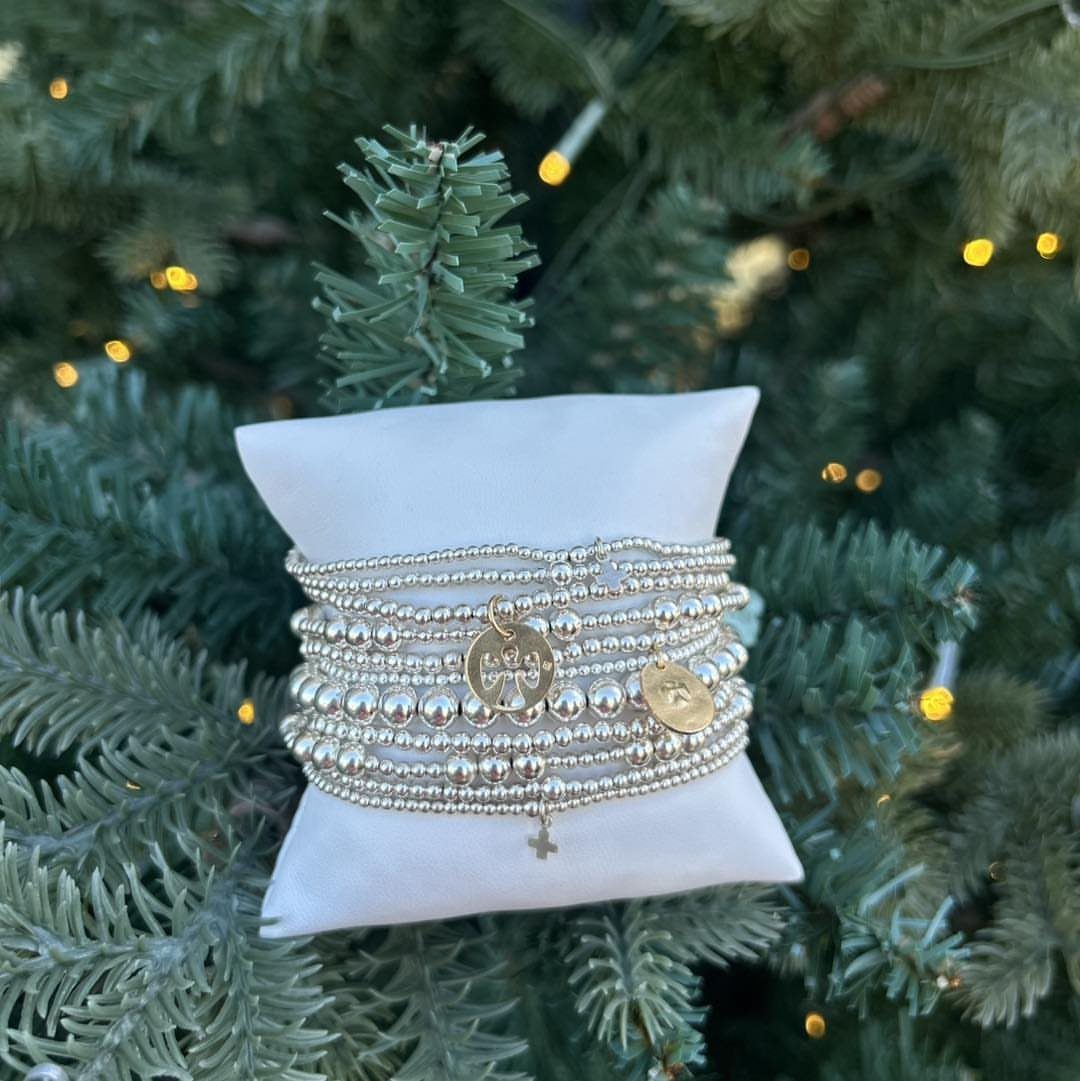 Silver beaded bracelets with small metal charms resting on a white pillow in evergreen branches.