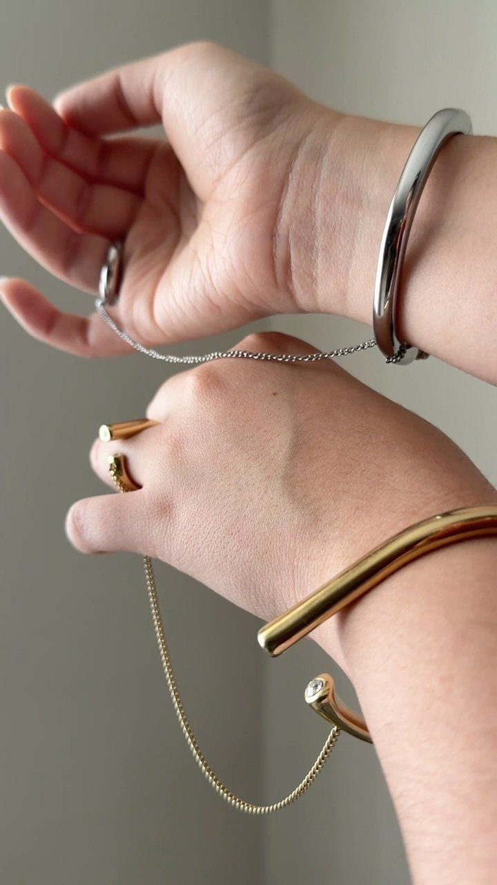 Hands wearing thick metal cuff bracelets and gold rings connected by a delicate chain.