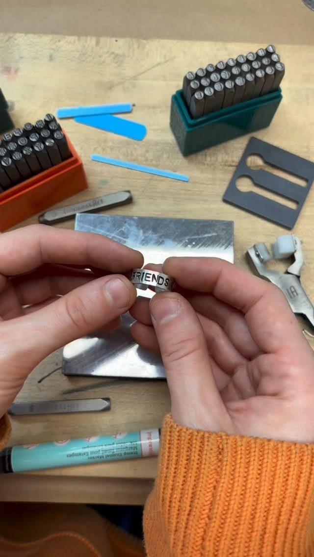Hands holding a small silver ring stamped with the word FRIENDS on a workbench.