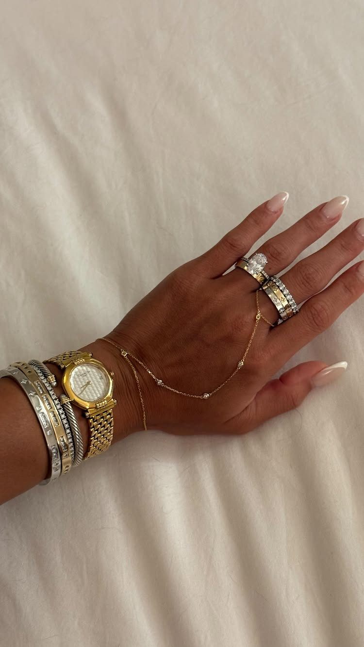 A hand adorned with stacked gold and silver bracelets, rings, a watch, and a delicate hand chain.