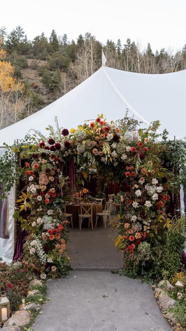 19 Mountain Wedding Ideas for Scenic, Cozy Celebrations 2 Floral arch with autumn leaves and red flowers leading into a white wedding tent.