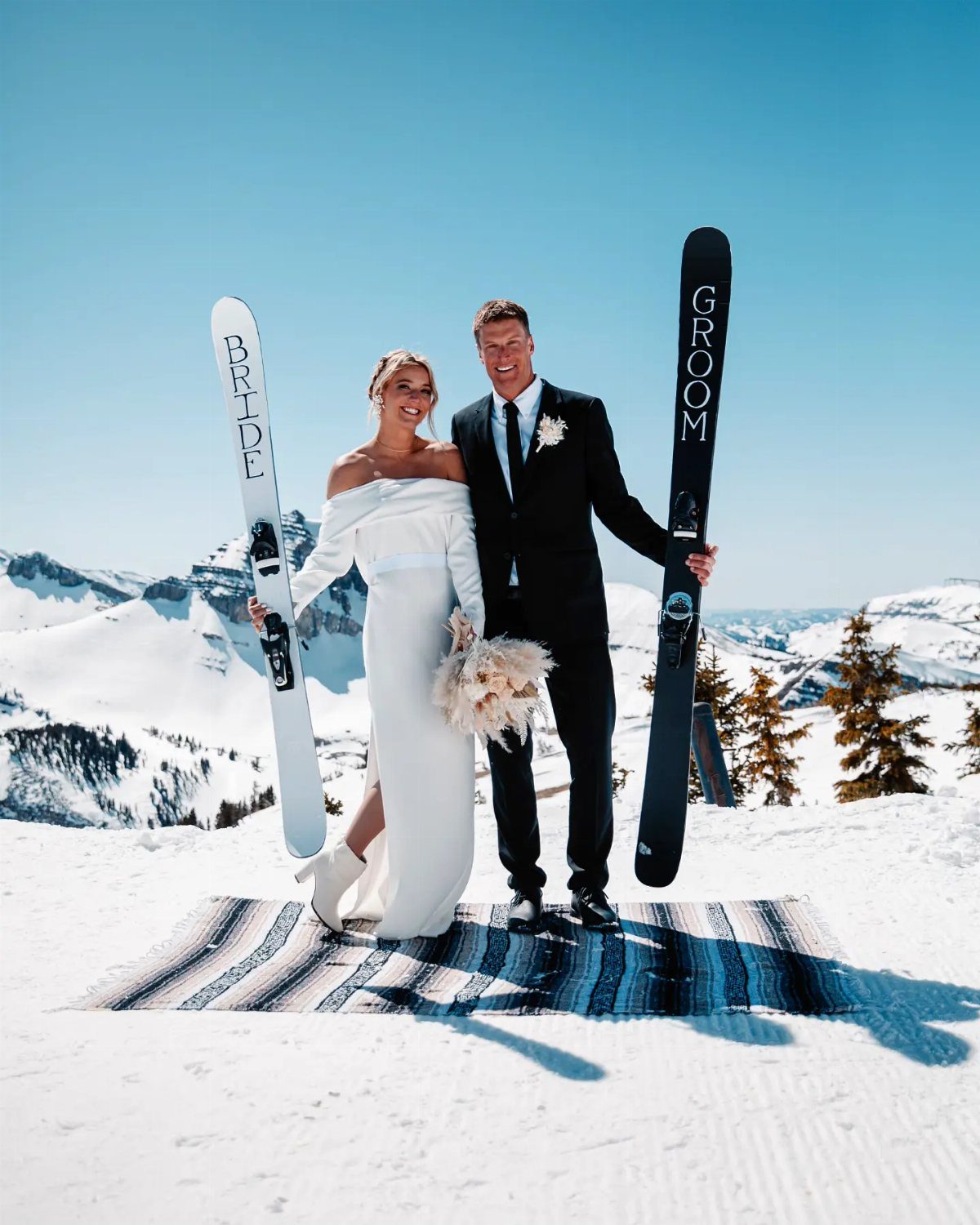19 Mountain Wedding Ideas for Scenic, Cozy Celebrations 5 Bride and groom in wedding attire posing with skis in a snowy mountain setting.