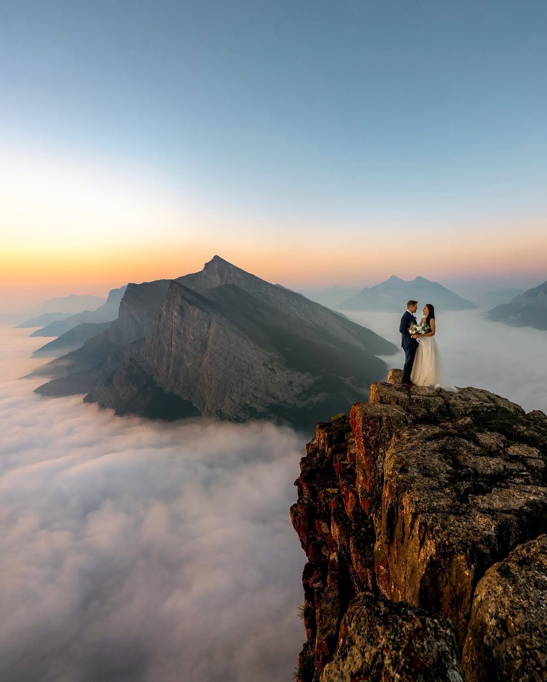 19 Mountain Wedding Ideas for Scenic, Cozy Celebrations 4 Bride and groom standing on a rocky cliff overlooking mountains at sunrise.