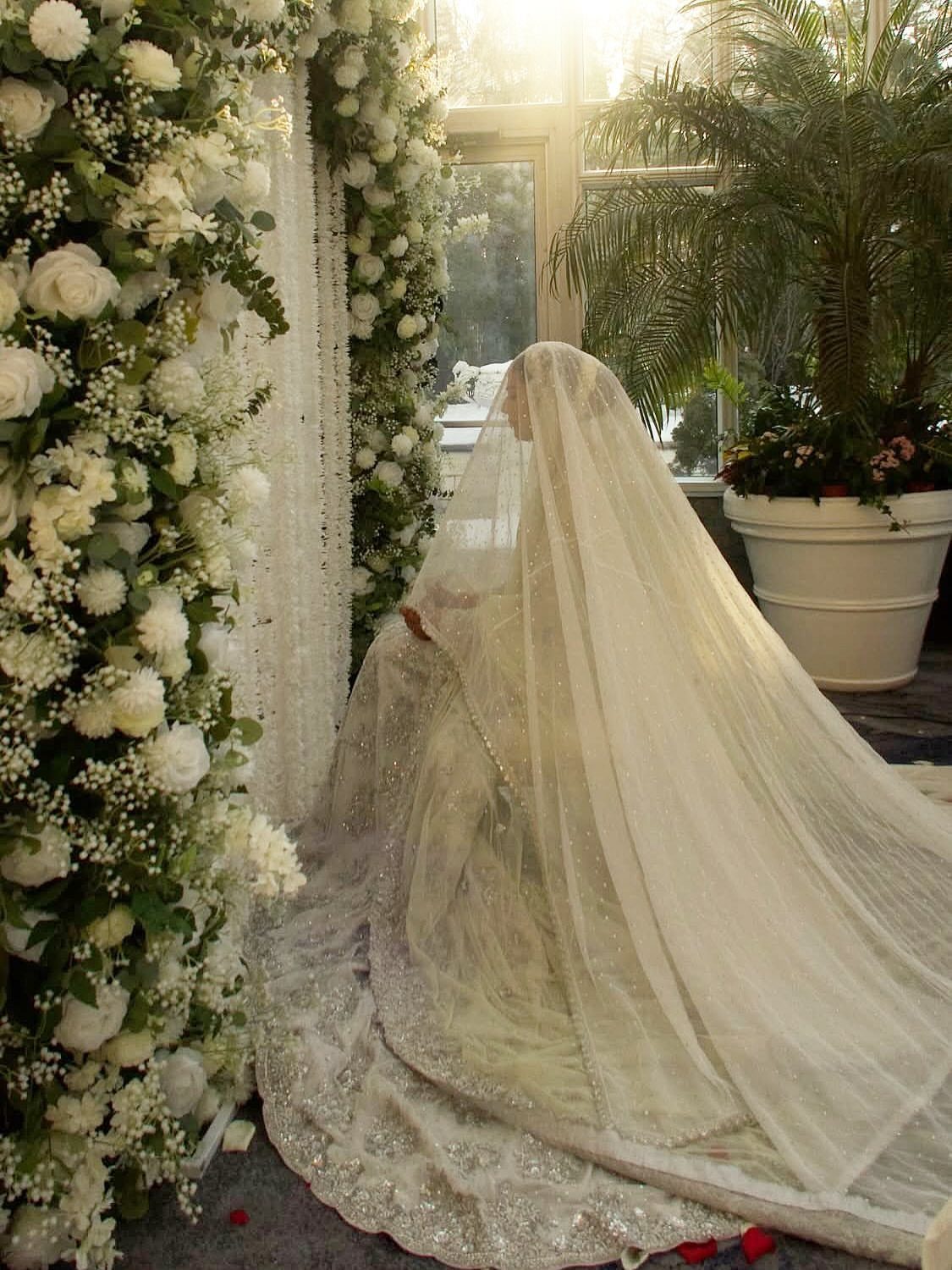 26 Nikah Dress Ideas for Your Perfect Wedding Ceremony 18 Bride in white gown and veil standing by a white floral wall.