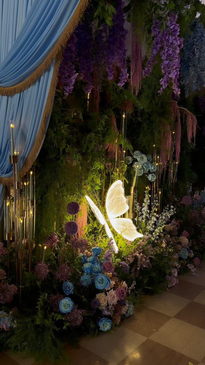 Glowing white butterfly sculpture nestled among purple and blue flowers and blue curtains.