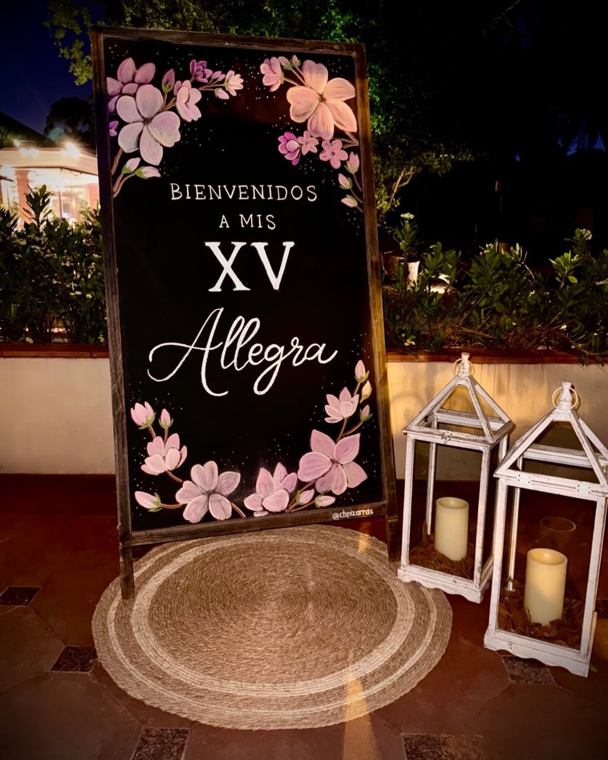 Chalkboard welcome sign with pink flowers and white lanterns on a jute rug.