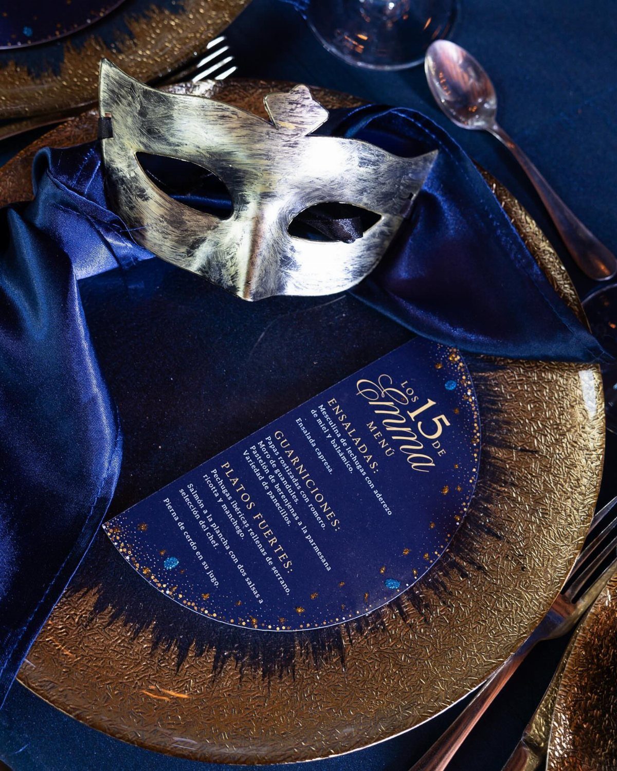 Silver masquerade mask on a blue satin napkin and gold plate with menu.