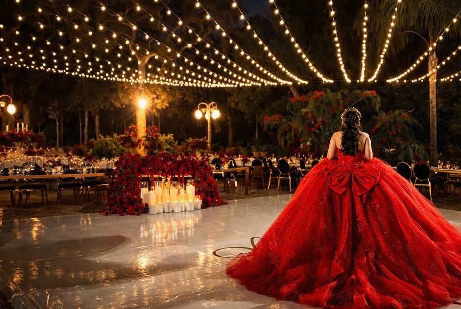 Woman in red ballgown facing an outdoor event with string lights and red flowers
