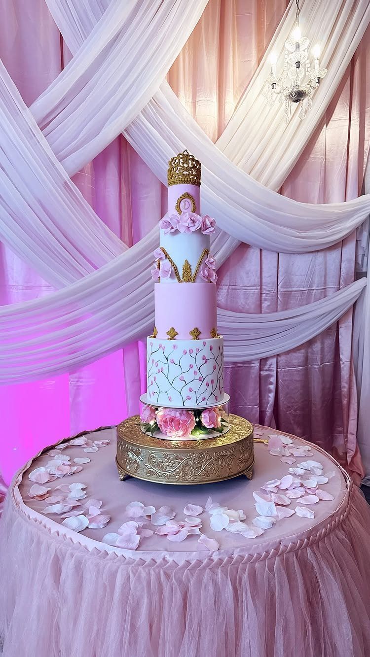 Multi-tiered pink and white cake with a gold crown on a decorated table.
