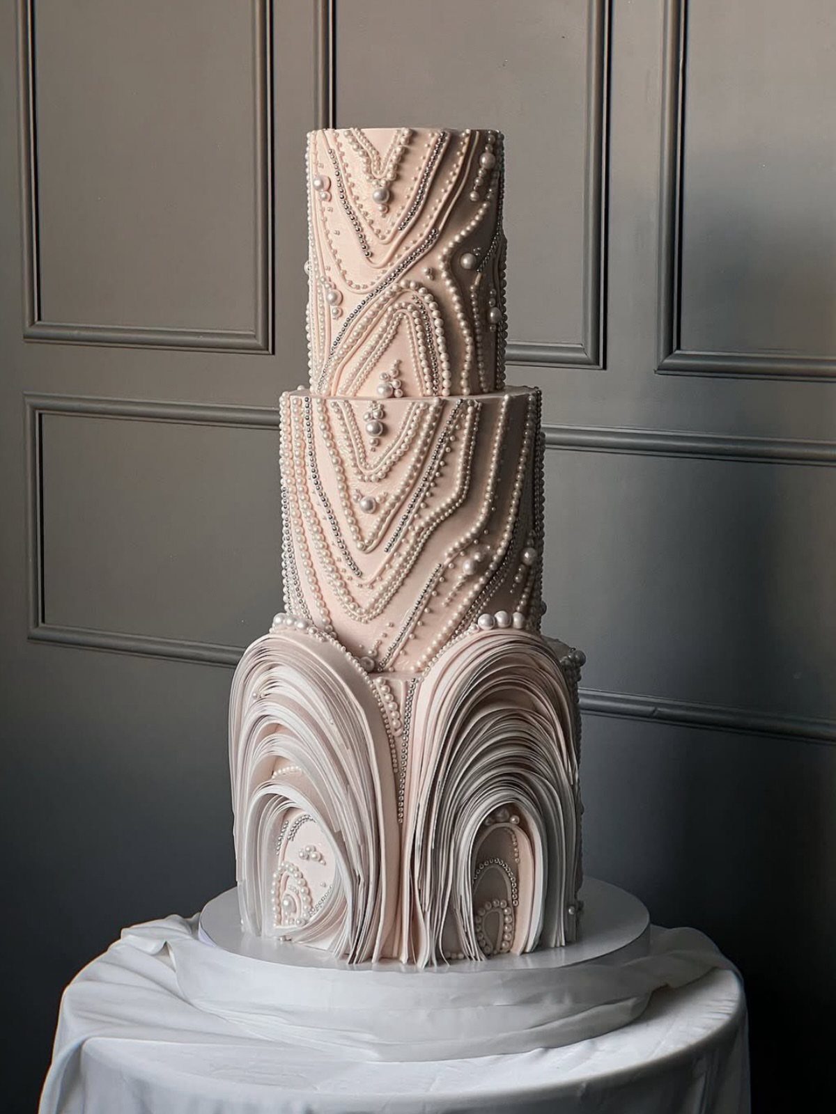 38 Romantic Wedding Cake Ideas for Your Celebration 23 Three-tier pale pink cake with pearl beading and large bottom ruffles against grey walls.