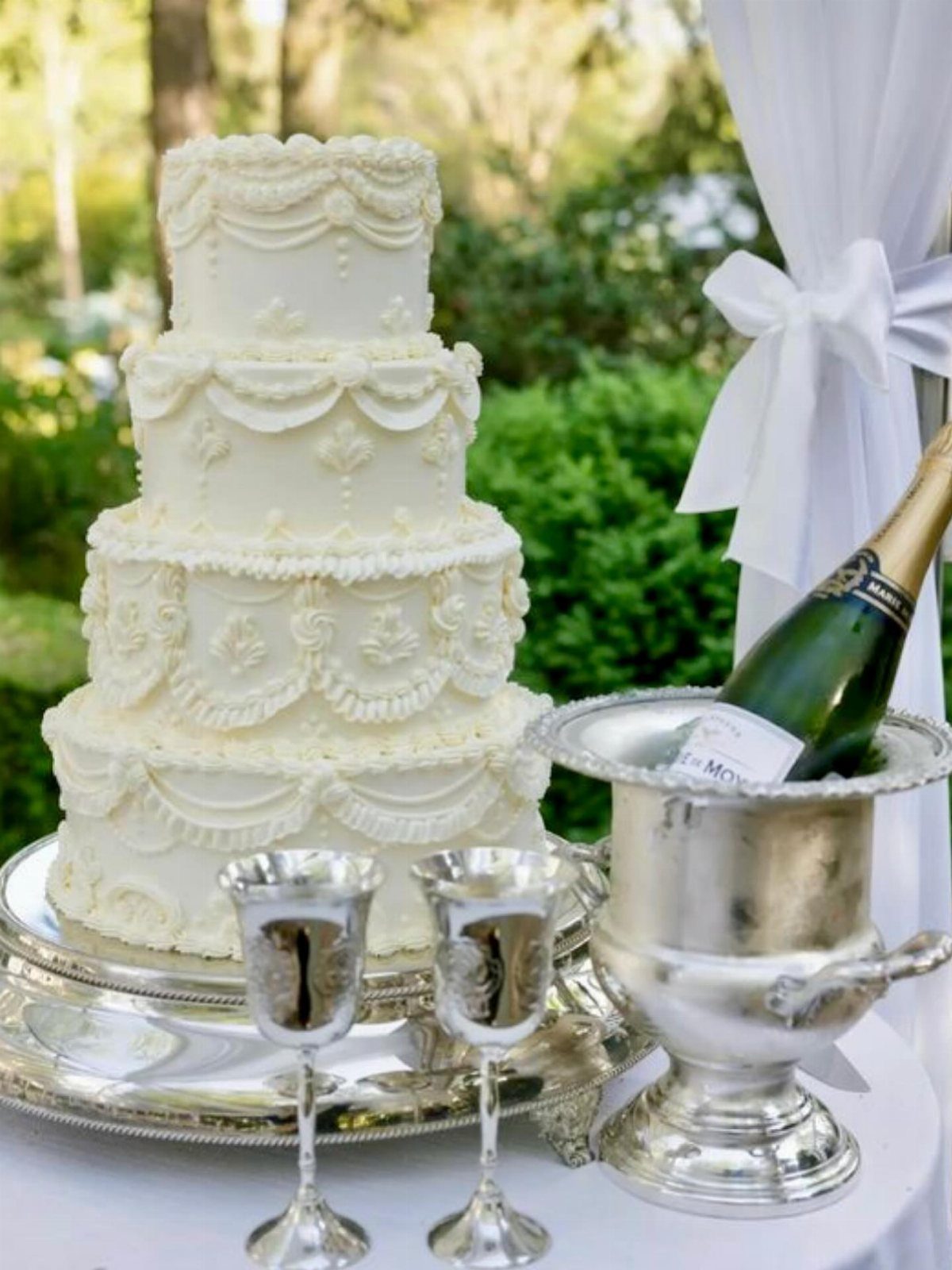 38 Romantic Wedding Cake Ideas for Your Celebration 20 Four-tier white wedding cake with ornate piping next to silver champagne bucket and goblets.