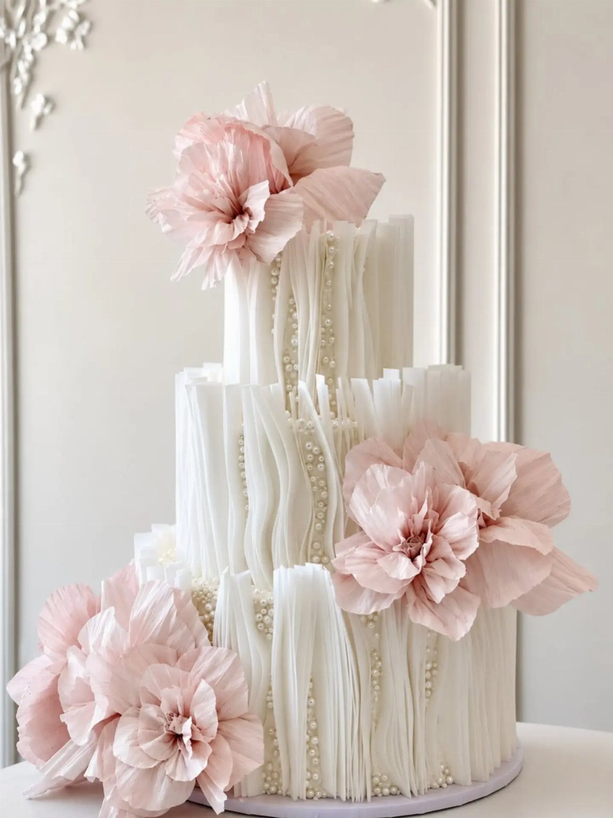 38 Romantic Wedding Cake Ideas for Your Celebration 28 Three-tier white wedding cake with pleated frosting and large pink wafer-paper flowers.