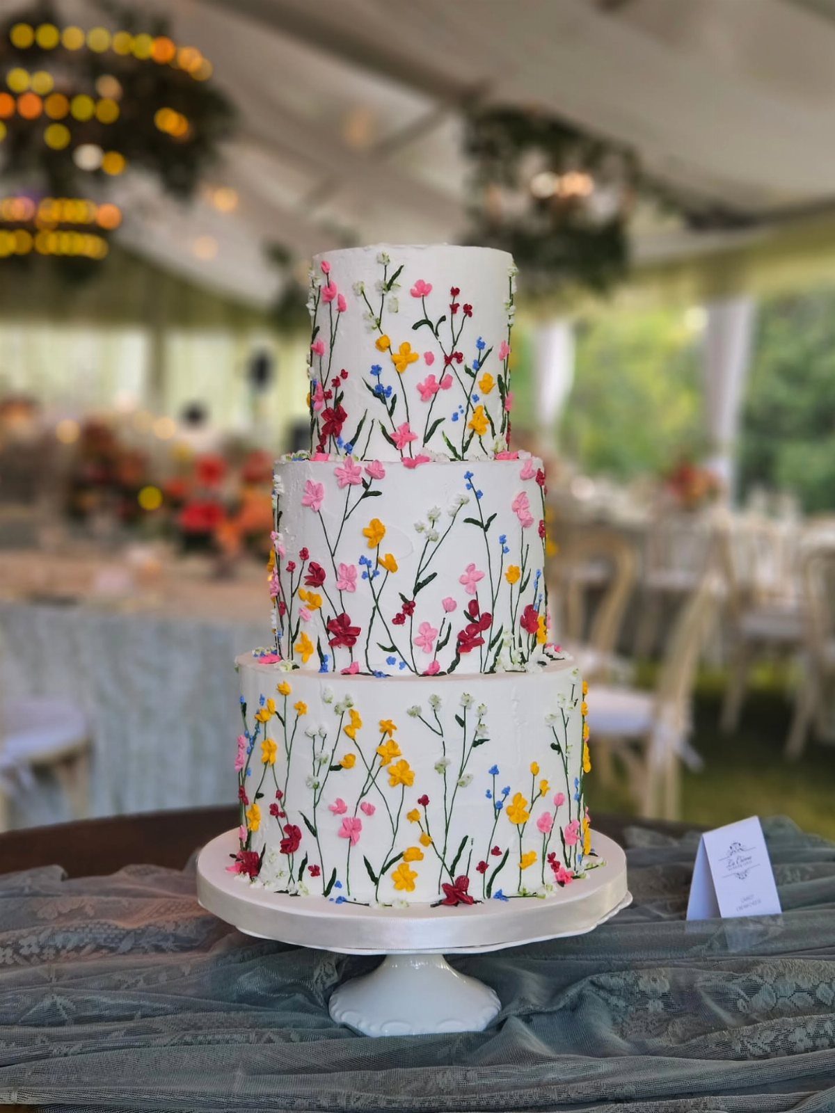38 Romantic Wedding Cake Ideas for Your Celebration 22 Three-tier white wedding cake decorated with colorful painted wildflowers on a white stand.