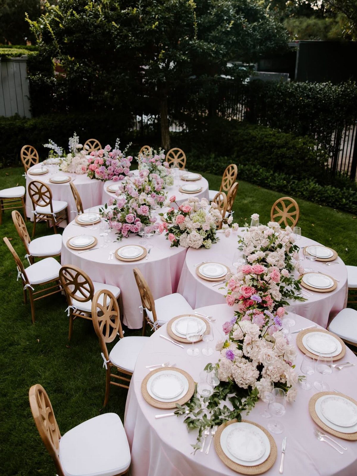 13 Round Table Wedding Decor Ideas: From S-Curved Layouts to Draped Florals 11 Round wedding tables with pink tablecloths and large pink and white floral centerpieces on a lawn.