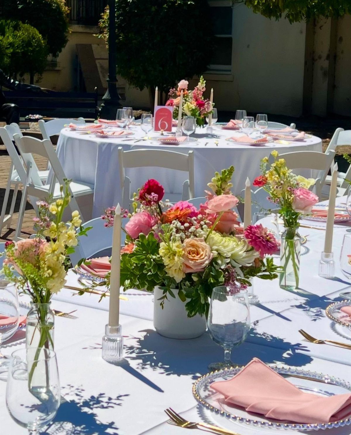 13 Round Table Wedding Decor Ideas: From S-Curved Layouts to Draped Florals 4 Floral arrangements and pink napkins set on white round wedding tables outdoors.