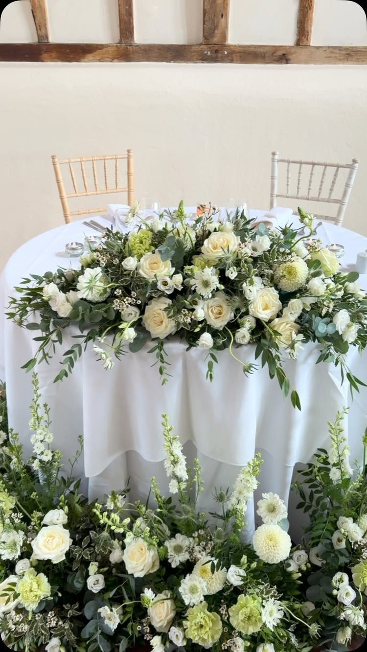 13 Round Table Wedding Decor Ideas: From S-Curved Layouts to Draped Florals 12 White floral arrangement draped over a round table with a white tablecloth and wooden chairs.