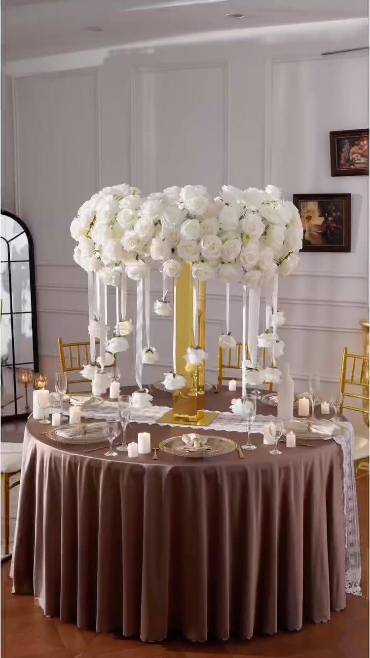 13 Round Table Wedding Decor Ideas: From S-Curved Layouts to Draped Florals 3 A tall gold pedestal holding a large arrangement of white roses with hanging ribbons on a brown tablecloth.
