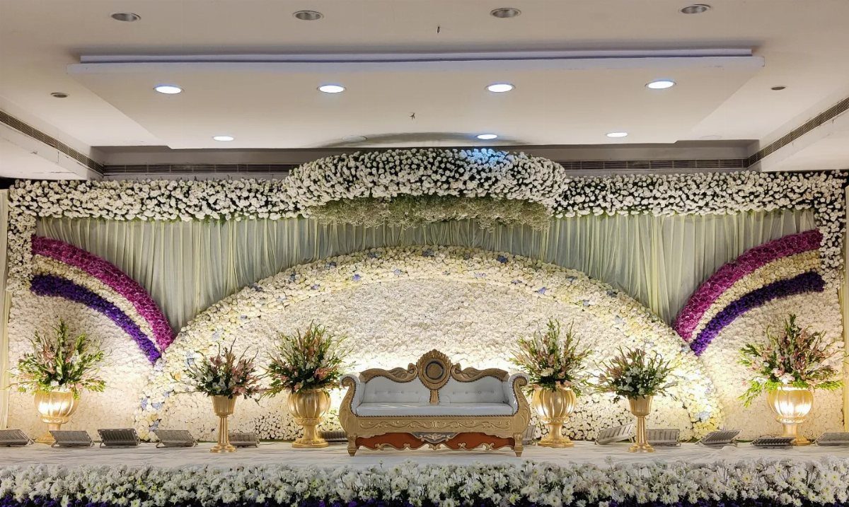 Wedding stage with white floral backdrop, purple fabric drapes, and a white sofa.