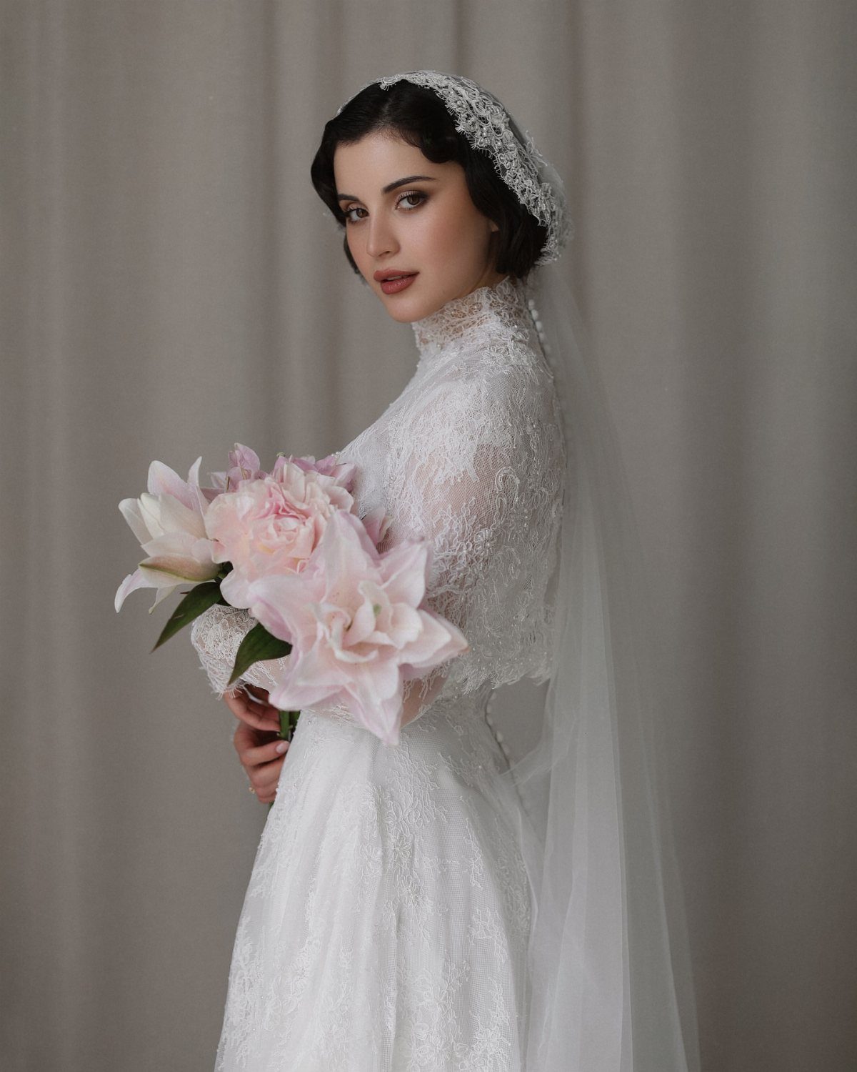 Woman in a white lace wedding dress holding pink lilies and wearing a long white veil.