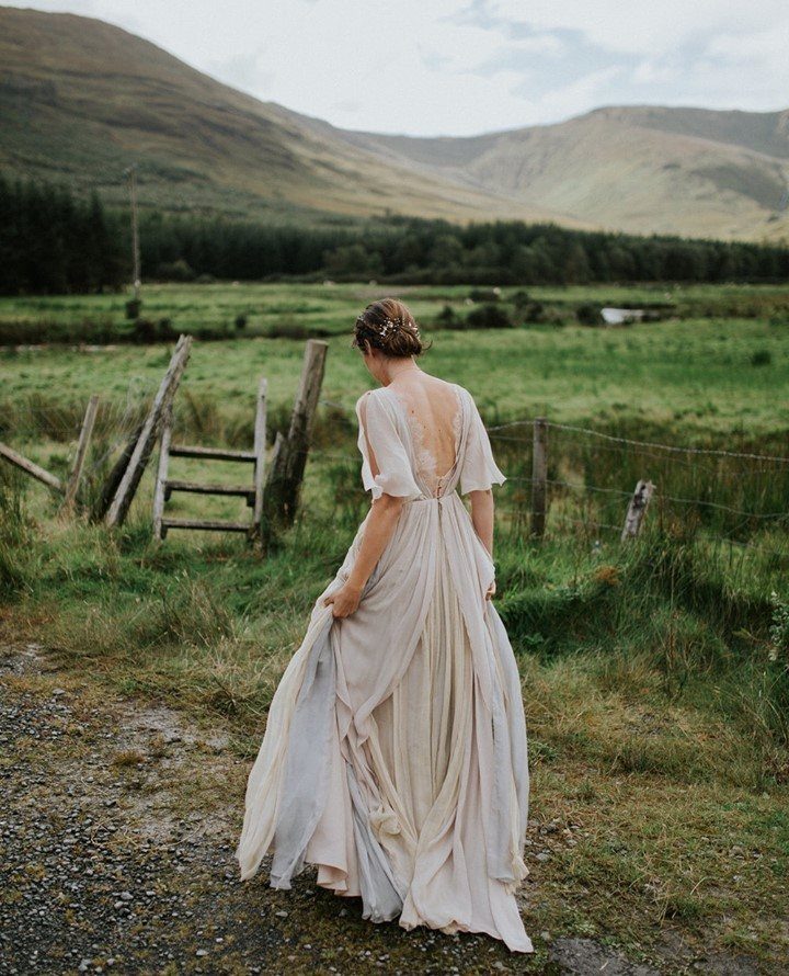 Woman in a flowing backless chiffon gown standing in a mountainous green landscape.