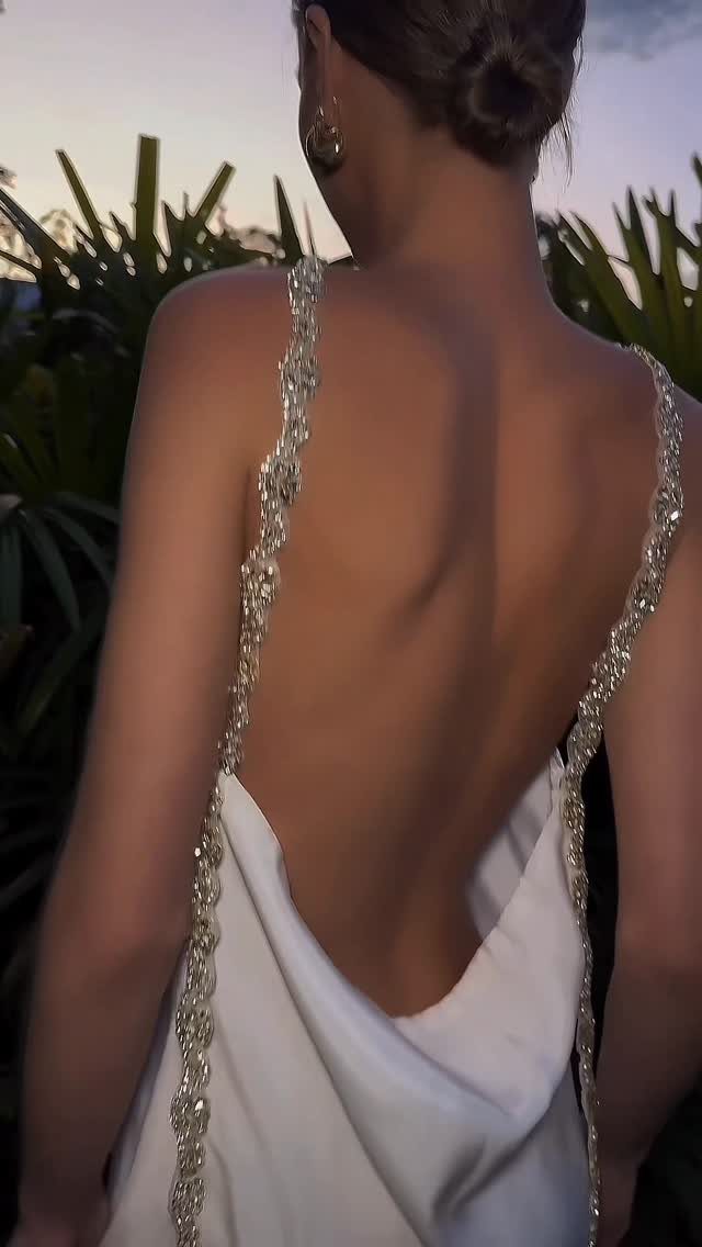Woman in a white backless gown with sparkling embellished straps against tropical greenery.