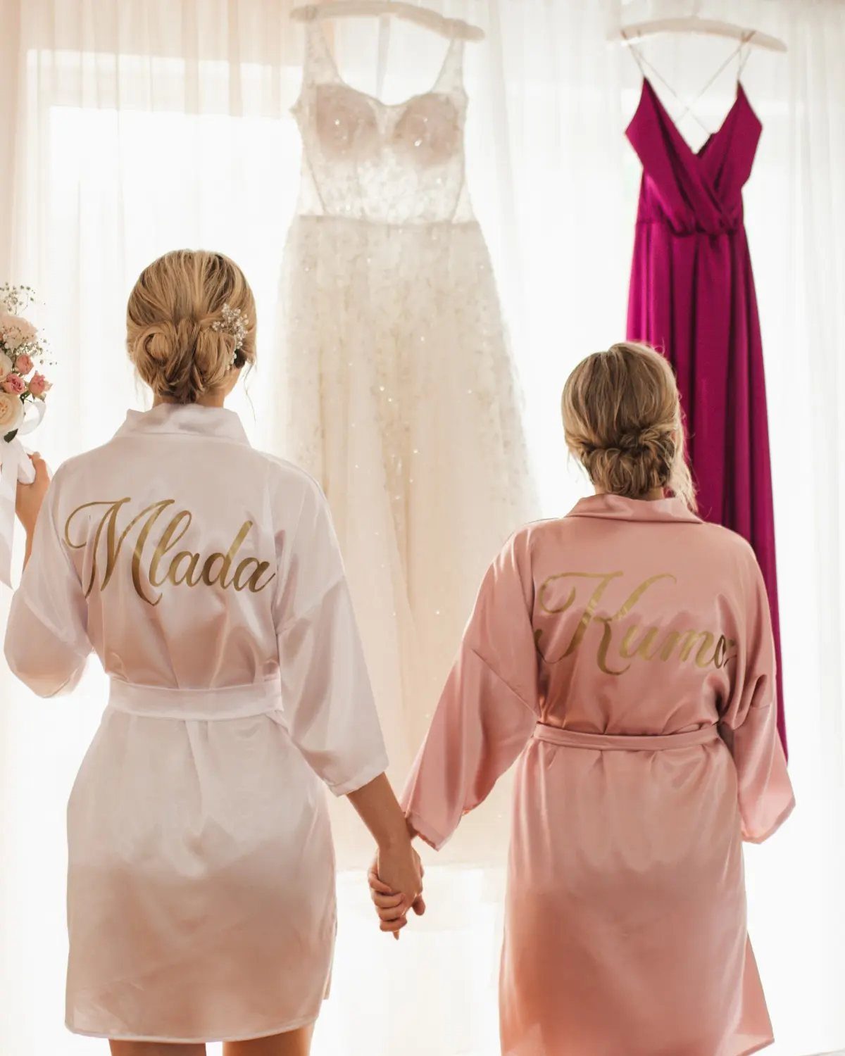 Sister Wedding Gift Ideas: 34 Picks She'll Treasure 8 Two women in personalized satin robes holding hands before a wedding dress.
