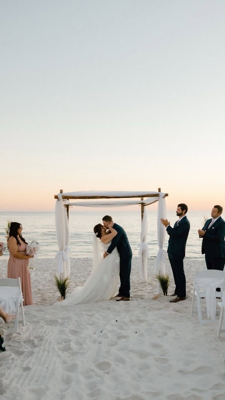 Bride and groom kissing under a white canopy on a sandy beach wedding.