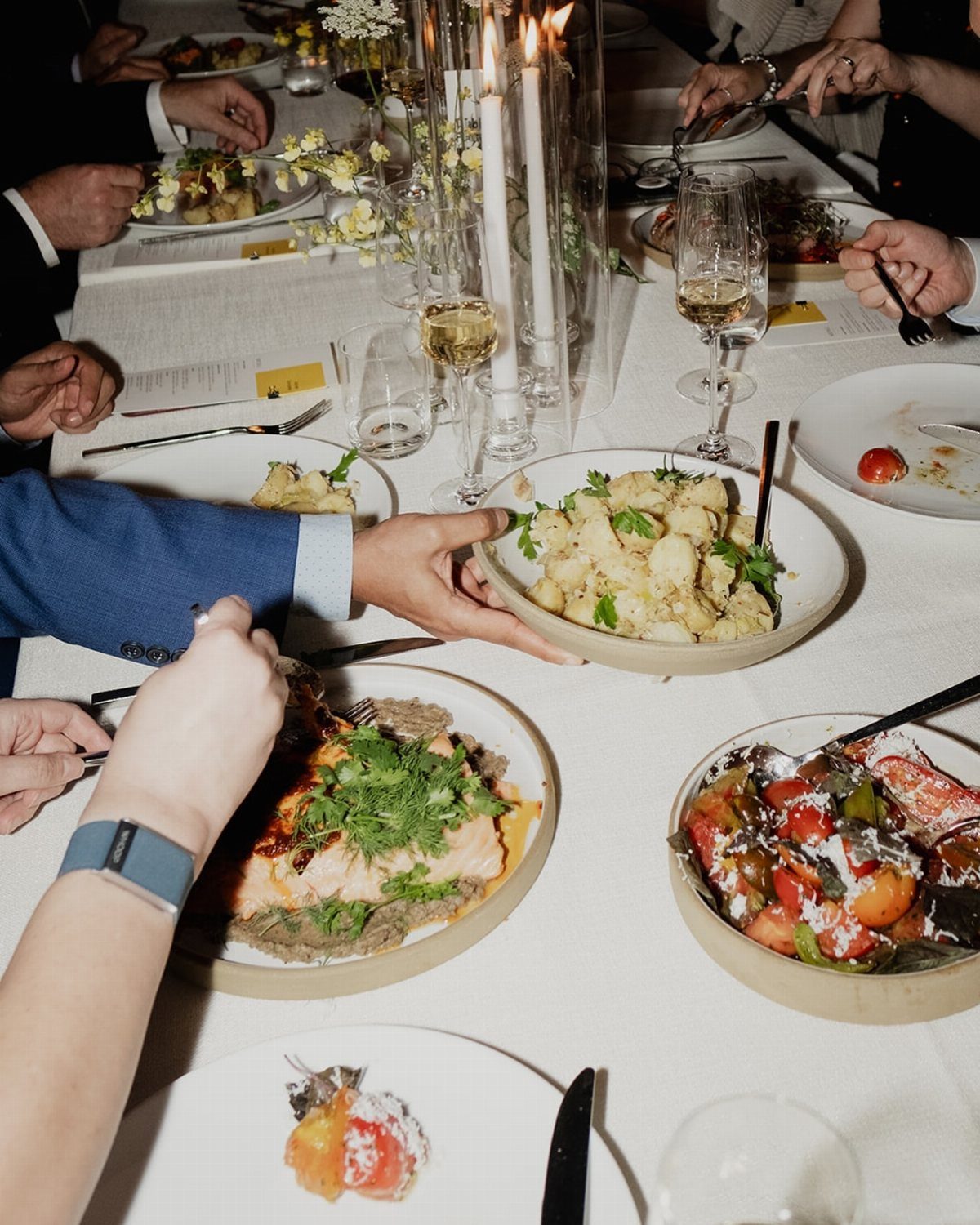 Shared bowls of food and wine glasses on a white table with candles.