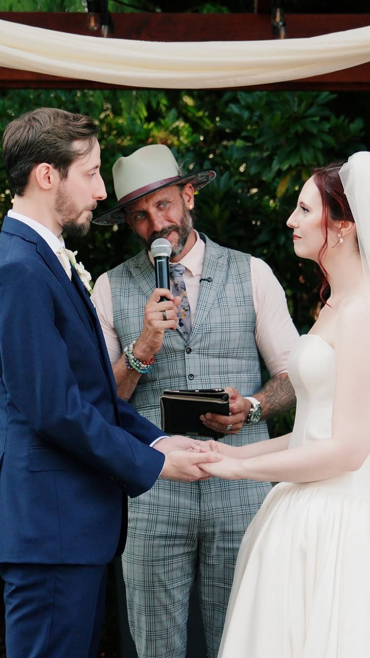 Wedding couple holding hands while a man in a hat officiates their ceremony.