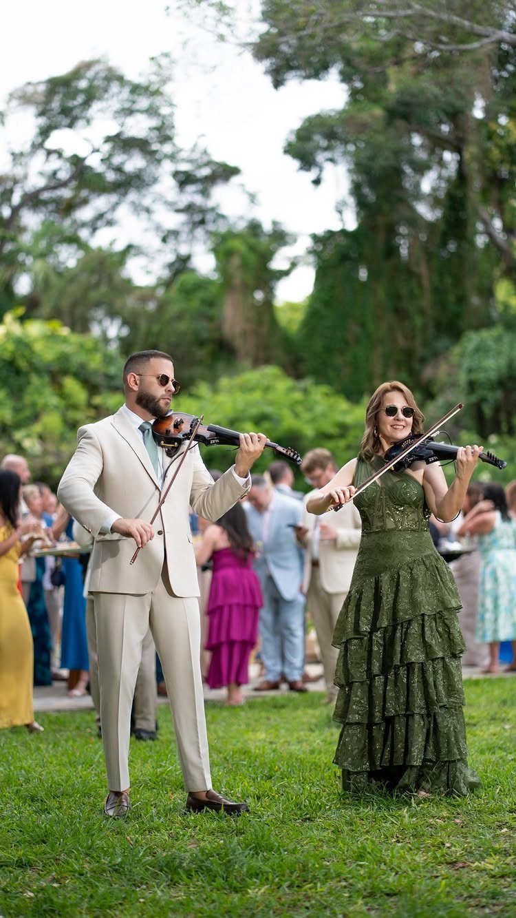 Two musicians playing violins on a green lawn at an outdoor wedding event.