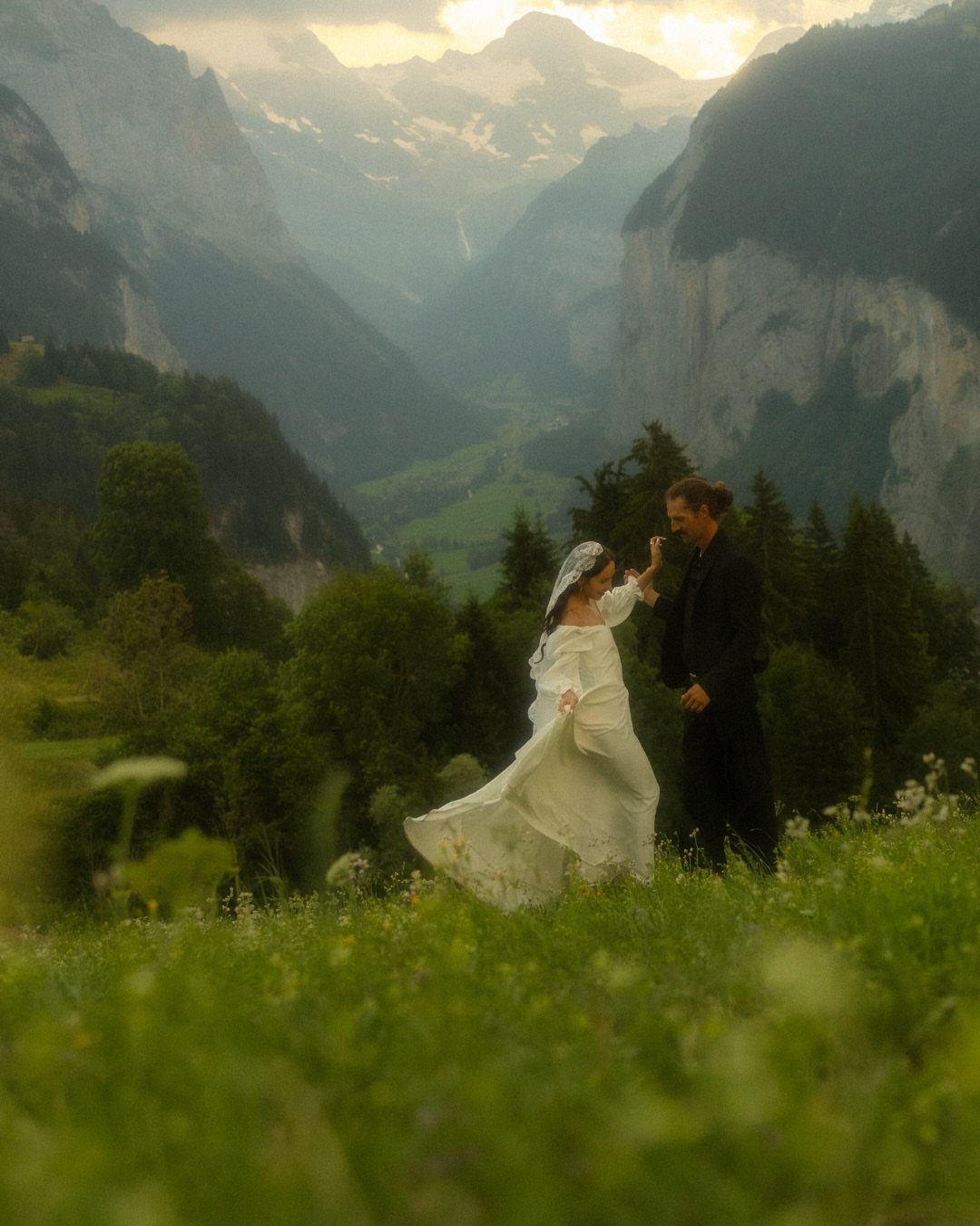 Bride and groom embracing in a grassy meadow overlooking a dramatic mountain valley.