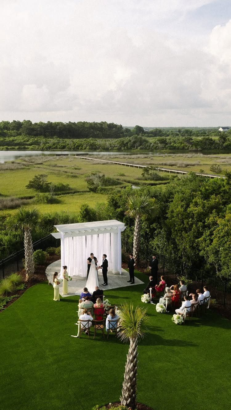 Wedding ceremony under a white pergola overlooking a coastal marsh and boardwalk.