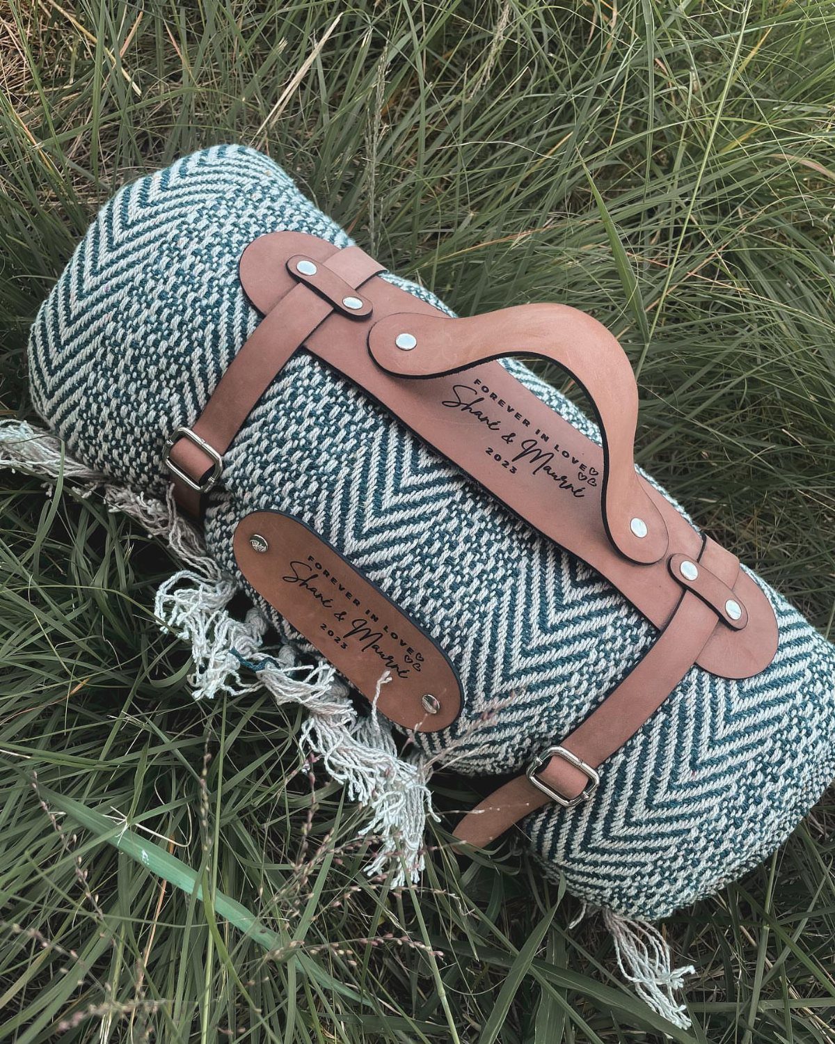 20 Small Wedding Gift Ideas for Couples Who Have Everything 7 Rolled herringbone wool blanket with a personalized engraved leather carrying strap on grass.