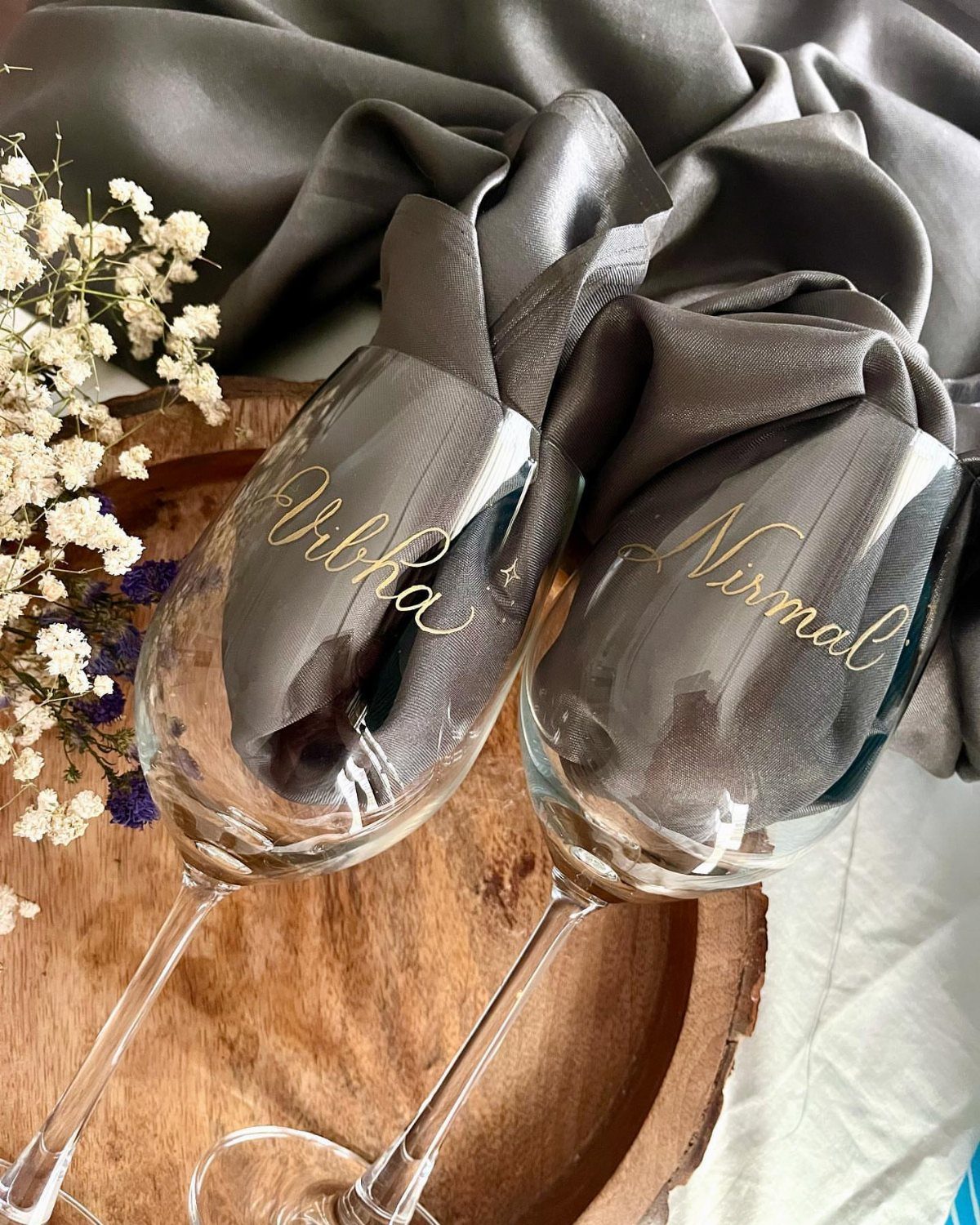 20 Small Wedding Gift Ideas for Couples Who Have Everything 5 Two personalized wine glasses with gold names on a wooden tray with flowers.