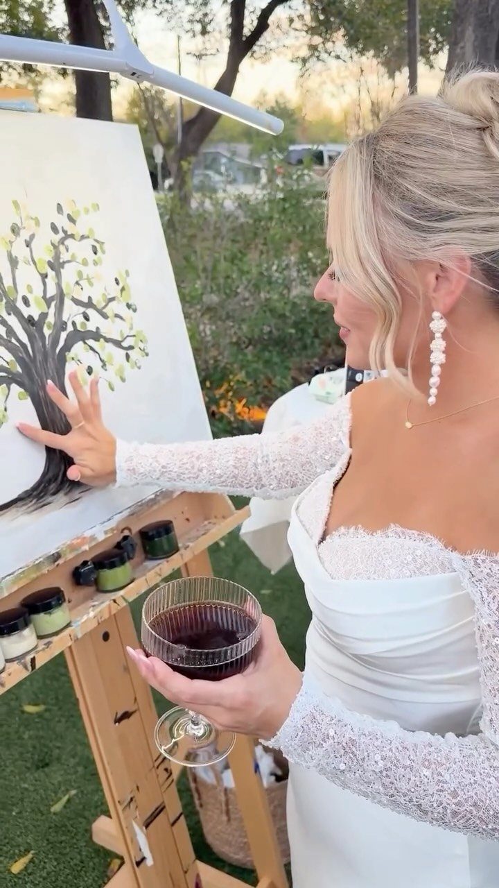 19 Wedding Activities for Guests: Reception Ideas Fun Enough to Fill the Whole Night 18 A bride in a lace dress painting a tree on an easel while holding wine.