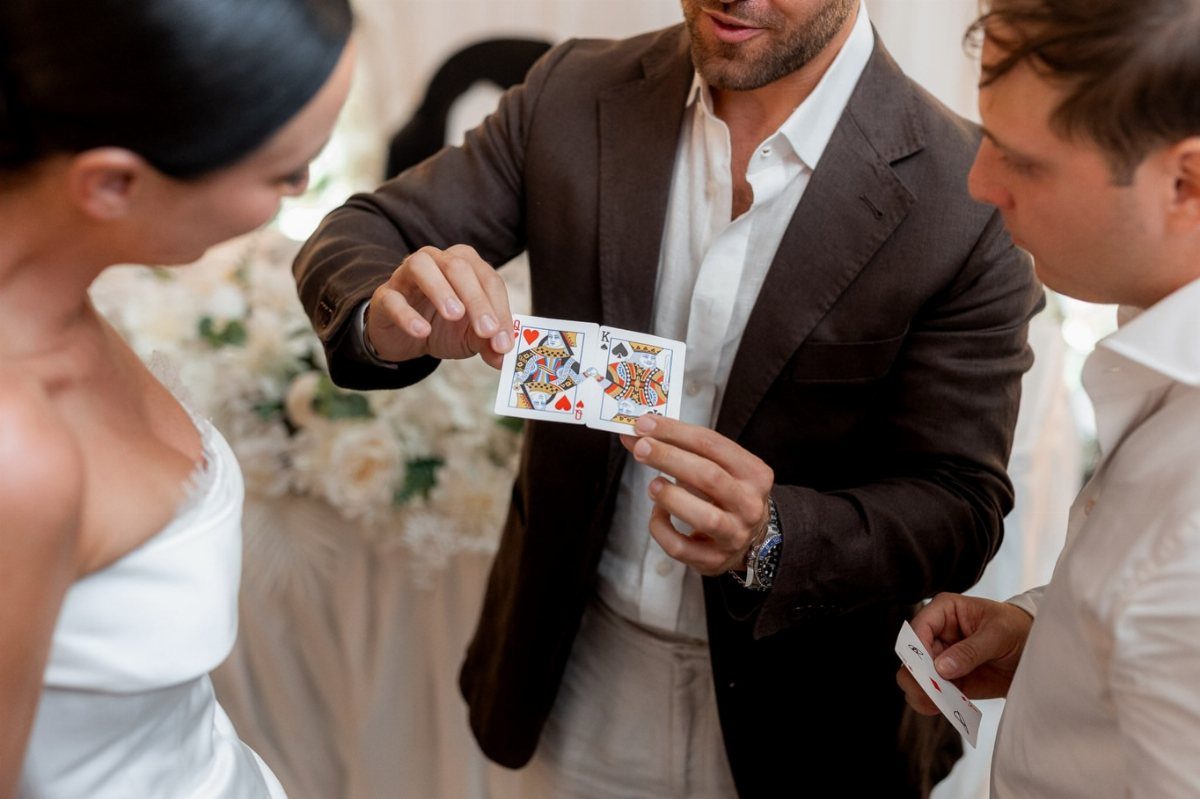 19 Wedding Activities for Guests: Reception Ideas Fun Enough to Fill the Whole Night 4 A man in a brown suit performs a card trick for a bride and a guest.