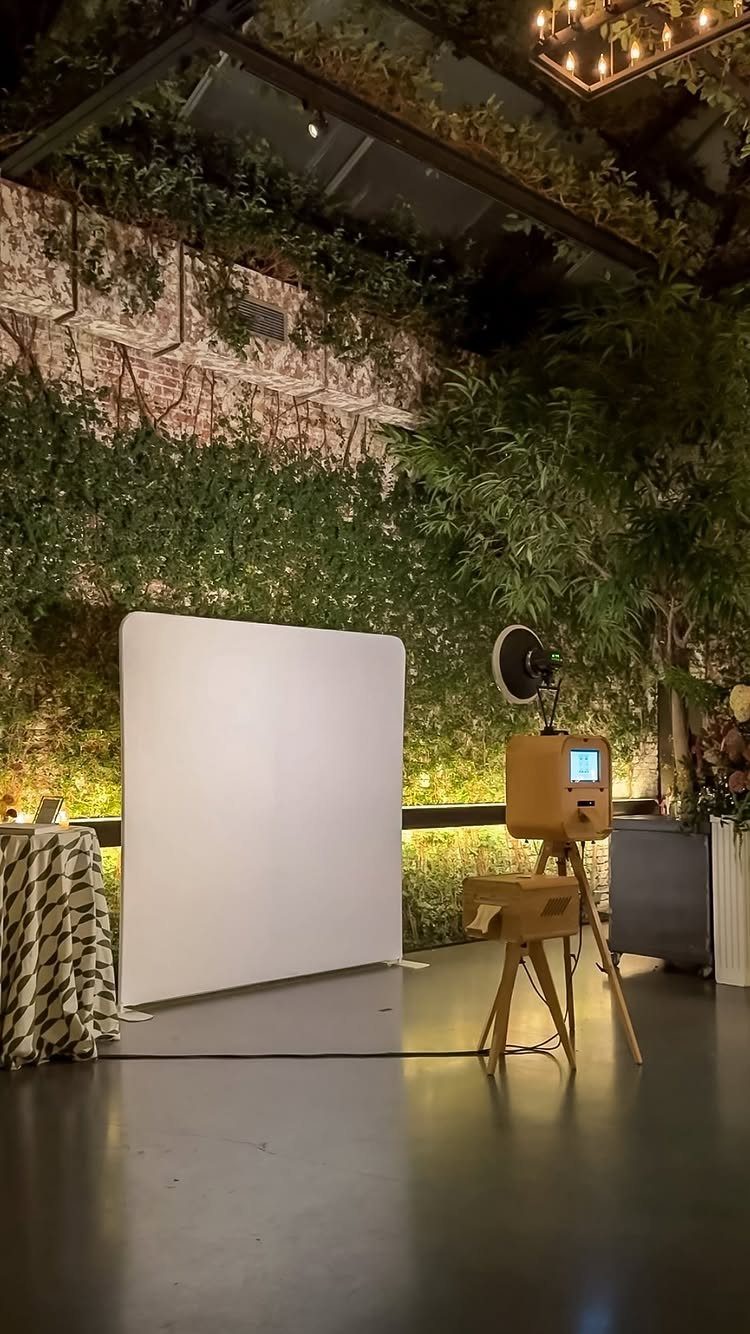 19 Wedding Activities for Guests: Reception Ideas Fun Enough to Fill the Whole Night 2 A wooden photo booth on a tripod positioned next to a large white backdrop in a lush venue.