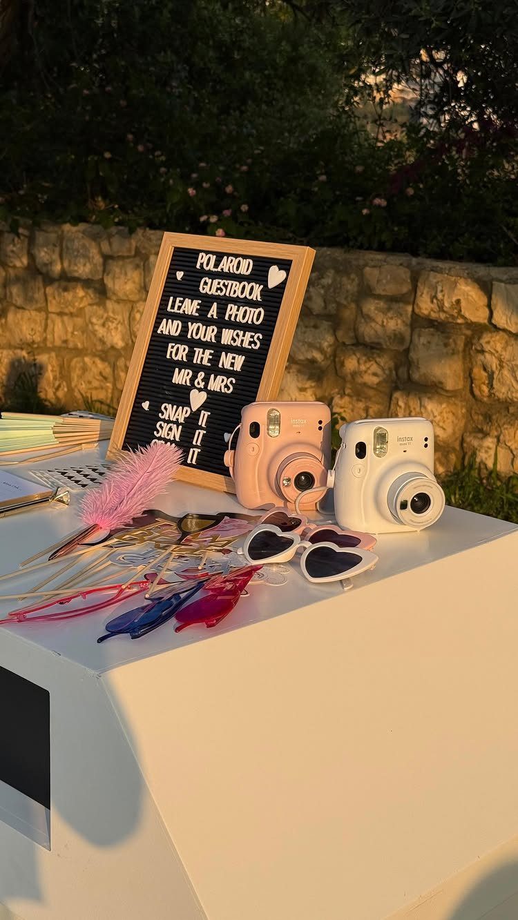 19 Wedding Activities for Guests: Reception Ideas Fun Enough to Fill the Whole Night 19 Polaroid guestbook station with two instant cameras, a letter board, and colorful novelty sunglasses on a white table.