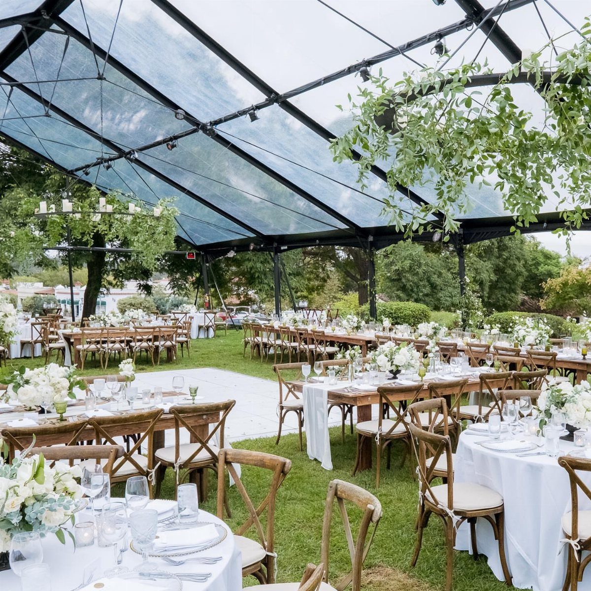 35 Backyard Wedding Reception Ideas for an Unforgettable Day 16 Clear-top wedding tent sheltering wooden dining tables and white flowers on a green lawn.