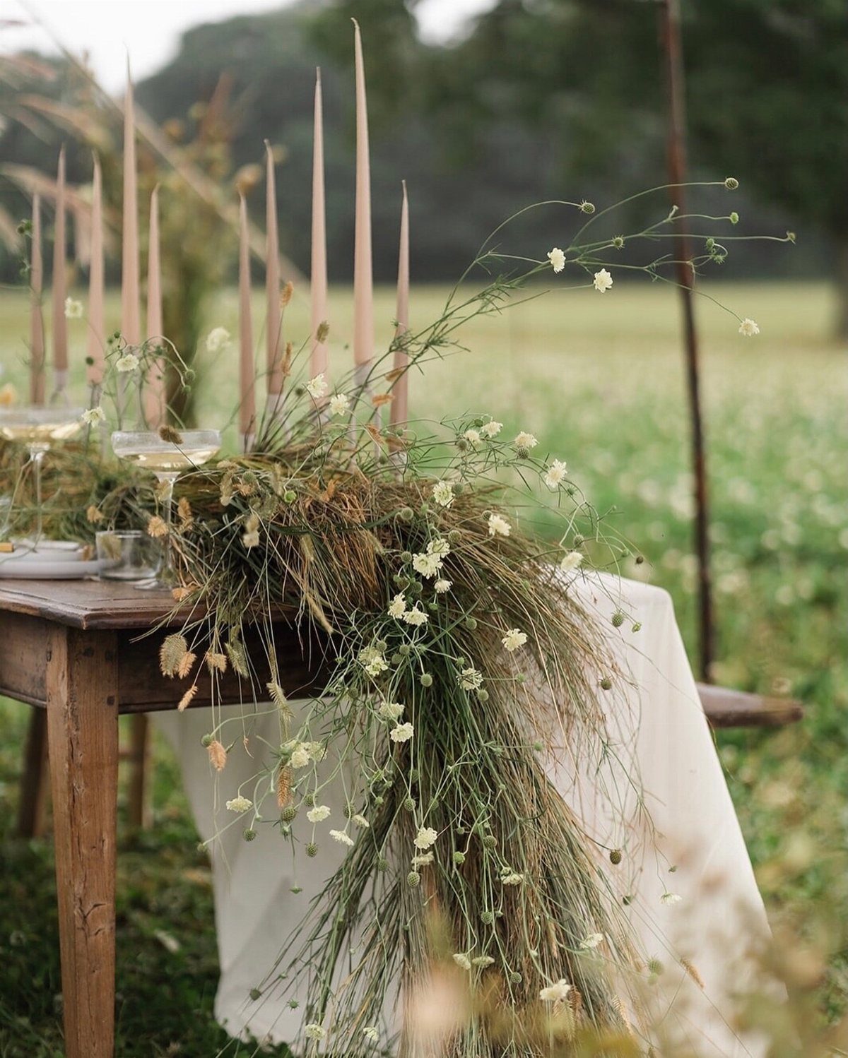 35 Backyard Wedding Reception Ideas for an Unforgettable Day 24 Rustic wooden table with dried flower arrangement and tall beige taper candles outdoors.